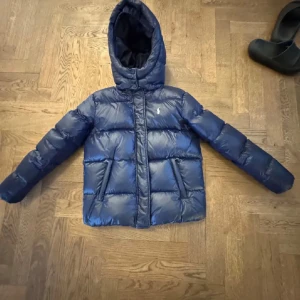 Blå dunjacka från Polo Ralph Lauren - Snygg blå dunjacka från Polo Ralph Lauren med huva och två fickor framtill. Jackan är quiltad och har dragkedja. Perfekt för kalla dagar och har en klassisk, sportig look. Märkets ikoniska logga broderad på bröstet. Pris kan diskuteras