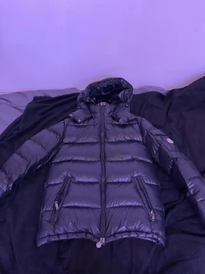 Svart Moncler pufferjacka med huva - Säljer en svart pufferjacka från Moncler med glansig finish och huva. Jackan har två framfickor med dragkedja, en ficka med knapp på ärmen och Moncler-logga. Fylld med dun och har quiltad design, perfekt för kalla dagar. 