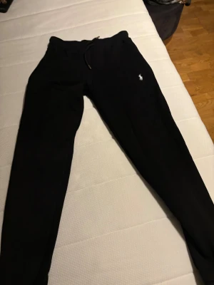 Svarta mjukisbyxor från Polo Ralph Lauren - Svarta mjukisbyxor från Polo Ralph Lauren med klassisk logga broderad i vitt på vänster ben. Byxorna har snörning i midjan och är tillverkade i mjukt bomullsmaterial. Perfekta för chill eller träning, med en skön loose passform.