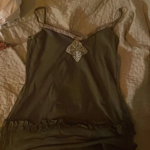 Olivgrön linnetopp med spetsdetalj - Säljer en olivgrön linnetopp från Temple i storlek S. Toppen har smala axelband, spetskant vid halsringningen och en dekorativ spetsapplikation framtill. Nedtill finns en volangdetalj som ger extra stilpoäng. Perfekt för dig som gillar unika detaljer och mjuka material.