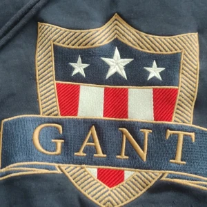 Mörkblå hoodie från GANT XL - Snygg mörkblå hoodie från GANT i storlek XL med stort broderat sköldmärke framtill i rött, vitt och blått. Tröjan har huva med snörning och ribbade muddar. Perfekt för dig som gillar klassisk och sportig stil. Köpt på Gant butiken för 1595 kr sparsamt använd 