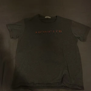 Mörkgrå Moncler t-shirt med logga - Snygg mörkgrå t-shirt från Moncler med tryckt logga i rött och svart på bröstet. Klassisk passform med rund hals och korta ärmar. Perfekt för dig som gillar stilrena och exklusiva plagg.