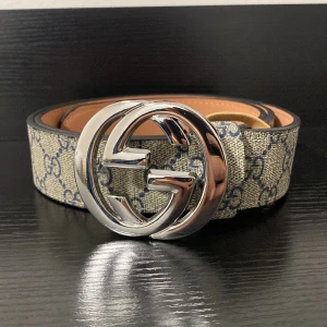 beige blått bälte - Säljer bälte med spänne i blank silverfärg. Bältet har beige och blått monogrammönster och insida i ljusbrunt skinn. Perfekt för dig som vill ha en lyxig och trendig accessoar med tydlig designerstil.