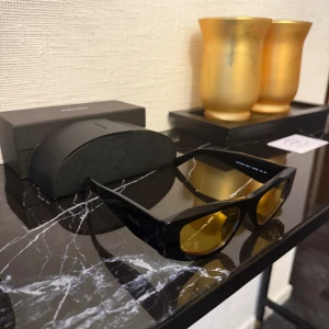 Svarta Prada solglasögon med gula linser - Säljer ett par snygga Prada solglasögon med bred, svart båge och coola gula linser. Glasögonen har en modern, kantig form och tydlig Prada-logga på sidan. Kommer med originalfodral och ask. Perfekt för dig som vill sticka ut med en unik look.