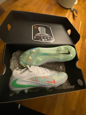 Nike Phantom GX EA Sports fotbollsskor - Nike Phantom GX EA Sports fotbollsskor med färgglad stickad ovandel i vitt, blått, grönt och rött. Snygga detaljer med grönt och blått Swoosh, samt genomskinlig plastsula med mönster och dobbar. Skorna har snörning och är designade för grym kontroll på planen. Köpta från unisportstore för 2745kr. Säljer för att dem är för små. + Helt nytt skick. Skickas med påse också.