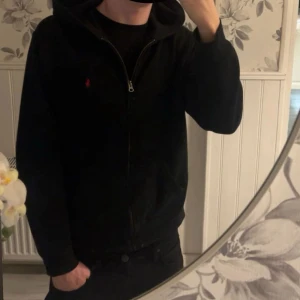 Svart hoodie från Polo Ralph Lauren XL - Svart hoodie från Polo Ralph Lauren i storlek XL. Klassisk design med huva, dragkedja framtill och rött broderat Polo-logo på bröstet. Perfekt för chill dagar och enkel att matcha med jeans eller joggers.