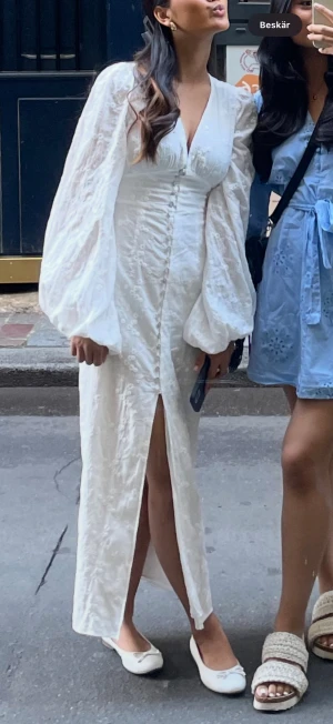 Vit långklänning med puffärmar - Säljer en elegant vit långklänning med v-ringning, puffiga långärmar och knappar framtill. Klänningen har en slits och är dekorerad med subtila broderade blommor i tyget. Perfekt för dig som vill sticka ut med en romantisk och trendig look.