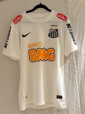 12/13 Vit Santos FC fotbollströja Nike - Santos FC matchtröja  i vit polyester med tryckta sponsorer i orange och svart. Klubbmärket med två stjärnor och årtal 1912-2012 på bröstet. Kortärmad modell med rund hals och ventilerande meshmaterial. Snygga detaljer på ärmarna och ryggen.