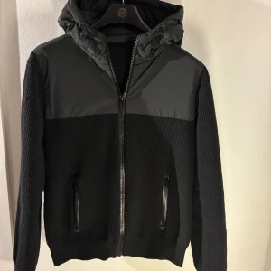 Ecoalf – Svart cardigan-jacka (stl L) - Säljer en svart cardigan-jacka från Ecoalf i storlek L. Den är dock liten i passformen och skulle bättre passa någon som normalt använder storlek M. Jackan har stickade ärmar och nederdel samt en lätt vadderad överdel och huva. Dragkedja framtill och två fickor med dragkedjor. Den är använd men i bra skick. Nypris är 2 499 kr.