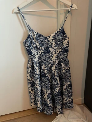 Blommig vit och blå kortklänning - Supersöt kortklänning med smala axelband och v-ringad urringning. Klänningen är vit med blått blommigt mönster och har en lätt utställd kjol. Materialet känns mjukt och luftigt, perfekt för varma dagar. Passformen är normal och den ger en fräsch och somrig vibe.