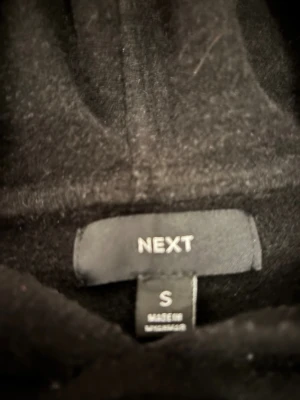 Svart hoodie från NEXT i storlek S - Svart hoodie från NEXT i storlek S. Tröjan har en klassisk huva och långärmade ärmar, med en enkel och stilren design. Perfekt för dig som gillar en clean look och vill ha något bekvämt att slänga på sig. Materialet känns mjukt och skönt mot huden.