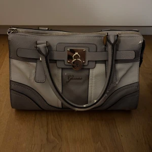 Grå handväska från Guess med guldiga detaljer - Snygg handväska från Guess i olika nyanser av grått med guldiga metalldetaljer och Guess-logga framtill. Väskan har dubbla handtag, dragkedja upptill och dekorativt lås. Tillverkad i syntetmaterial med stilren, strukturerad form. Pris kan diskuteras.