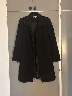 Svart kappa från WE Women, strl 40 - Svart kappa från WE Women i klassisk rak modell med krage och knappar framtill. Långärmad och tillverkad i mjukt ullmaterial som håller dig varm under kyliga dagar. Perfekt för dig som gillar stilrena och tidlösa plagg.