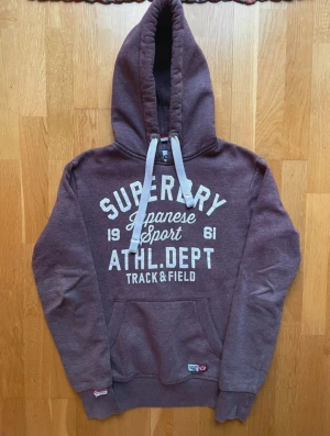 Vinröd hoodie från Superdry - Säljer en vinröd hoodie från Superdry med vit tryckt text på bröstet och stora snören i huvan. Tröjan har en klassisk känguruficka och ribbade muddar vid ärmslut och nederkant. Perfekt för chill dagar och sportiga outfits.