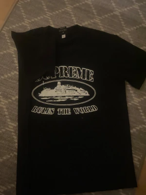 Cortiez svart t-shirt med tryck - Svart t-shirt från Cortiez med stort vitt tryck på bröstet som visar ett skepp och texten 'SUPREME RULES THE WORLD'. Klassisk rund hals och korta ärmar. Materialet är mjuk bomull som känns skönt mot huden. Perfekt för dig som gillar streetwear och statement-plagg. Pris kan diskuteras 