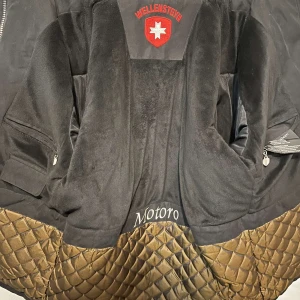 Svart parkajacka från Wellensteyn - Parkajacka från Wellensteyn i svart med quiltad insida och flera praktiska fickor framtill. Jackan har en tydlig logga på bröstet och ärmarna, samt grov dragkedja och knappar. Materialet är slitstark polyester och jackan har lång ärm och normal passform. Saknar luvan. 