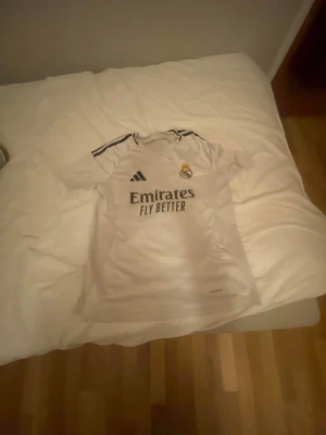 Real Madrid Mbappé 9 vit matchtröja - Säljer en vit Real Madrid fotbollströja från Adidas med Mbappé och nummer 9 på ryggen. Tröjan har svarta detaljer, klubbmärke på bröstet och klassiska Adidas-ränder på axlarna. Materialet är lätt och andas, perfekt för träning eller match. Äkta direkt från spaninen