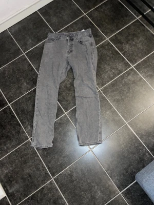 Grå jeans från Sweet SKTBS, stl 29 - Säljer ett par grå jeans från Sweet SKTBS med klassisk femficksdesign och rak passform. Jeansen har en enkel look med svarta sömmar och en snygg läderpatch bak i midjan. Perfekt för dig som gillar streetwear och skandinavisk stil.