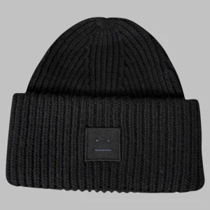 Svart ribbstickad mössa från Acne Studios - Svart ribbstickad mössa från Acne Studios med klassisk uppvikt kant och det ikoniska Face-logotypen framtill. Tjockt och mjukt material som håller dig varm och ger en clean look. Perfekt accessoar för kalla dagar.
