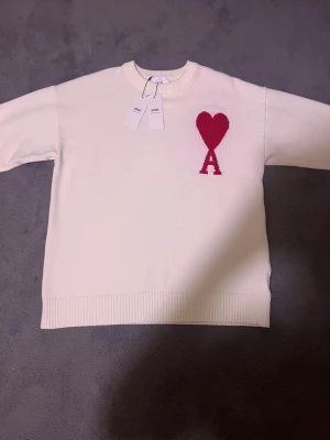 Vit stickad tröja från Ami Paris - Vit stickad tröja från Ami Paris med röd hjärt- och A-logga på bröstet. Rund hals och ribbade muddar vid ärmslut och nederkant plagget inkluderars med tags 🏷️ på tröjan och tröjan är Helt ny + 100% Bomull + 0 Defekter 