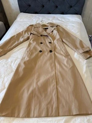 Beige trenchcoat från Vero Moda - Snygg beige trenchcoat från Vero Moda med klassisk dubbelknäppning, långa ärmar och bälte i midjan. Jackan har två fickor fram och är tillverkad i ett slätt, lätt material som ger en clean och stilren look. Perfekt för dig som gillar tidlös stil. Köpte för 800 aldrig använt 