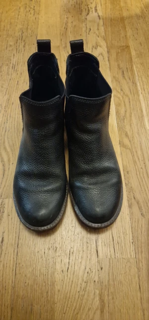 Svarta Ecco boots i skinn - Snygga svarta Ecco boots med rund tå och platt sula. Tillverkade i grovt skinn med elastiska paneler på sidorna för enkel på- och avtagning. Perfekta för dig som gillar klassisk stil med en modern touch.