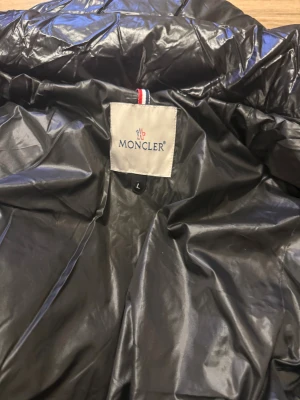 Svart Moncler pufferjacka med huva - Svart pufferjacka från Moncler med glansig finish, huva och coolt Moncler-märke på bröstet. Jackan har dragkedja med röd, vit och blå kant, praktiska fickor och ett serietidningsmönster på insidan. Perfekt för dig som vill ha en snygg och varm jacka.