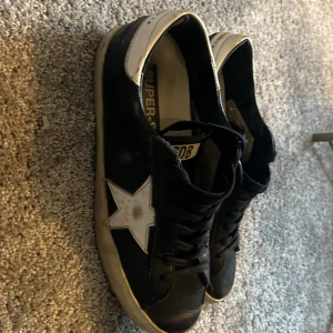 Golden Goose svartvita sneakers - Svarta sneakers från Golden Goose med vit stjärndetalj på sidan och vit häl. Klassisk låg modell med snörning och platt sula. Skorna är tillverkade i skinn och har en cool, urban vibe med kontrasterande detaljer. Nypris 5500