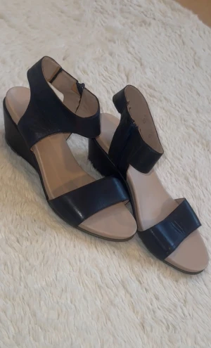 Mörkblåa kilklack-sandaler från Footwear - Snygga mörkblåa sandaler med kilklack från Footwear. Skorna har breda remmar i skinn, öppen tå och dragkedja på sidan för enkel påtagning. Innersulan är beige och mjuk mot foten. Perfekta för dig som vill ha både stil och komfort under sommaren.