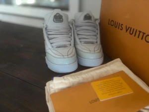 Louis Vuitton skor - Säljer ett par exklusiva Louis Vuitton sneakers i vitt skinn med rund tå och platt sula. Skorna har snörning, detaljerad LV-logga på plösen och en stor blomdetalj på sidan. Snygg textur på materialet och diskreta grå inslag på sulan. Perfekt för dig som gillar lyxiga streetwear-vibes.