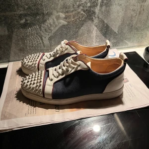 Christian Louboutin sneakers med nitar - Säljer ett par blå och vita Christian Louboutin sneakers med ikoniska röda sulor och silverfärgade nitar på tån. Skorna har snörning, rund tå och platt sula. Materialet är en mix av skinn och textil, med röda detaljer längs kanterna. Box och dustbags medföljer. Kvitto finns