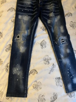 Dsquared2 blå jeans med slitningar - Snygga blå jeans från Dsquared2 med coola slitningar och hål på knäna, samt vita färgstänk för en edgy look. Jeansen har klassisk femficksdesign, knappgylf och tydliga tvättade detaljer. Passar dig som gillar streetstyle och vill sticka ut.