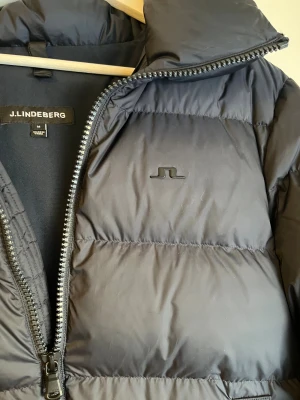 Svart pufferjacka från J.Lindeberg - Knappt använd! Mörk blå pufferjacka från J.Lindeberg i storlek M. Jackan har en rejäl huva, breda quiltade sektioner och två sidofickor. Materialet är syntet och jackan stängs med en grov dragkedja framtill. Perfekt för kalla dagar med sin fluffiga och stilrena look. (Pris kan diskuteras, kom med förslag!). INGA TÄKEN PÅ ANVÄNDNING!