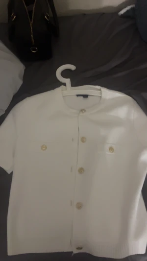 Vit kortärmad kofta från Zara - Vit kortärmad kofta från Zara med snygga guldfärgade knappar framtill och på fickorna. Tröjan har rund hals och är stickad i mjukt material. Perfekt för dig som gillar stilrena och klassiska plagg med en touch av lyx.