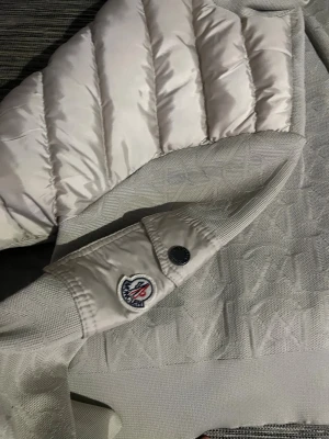 Beige pufferjacka från Moncler - Snygg beige pufferjacka från Moncler med quiltad framsida och stickade ärmar med diskret mönster. Jackan har svarta dragkedjor, ribbade muddar och en Moncler-logga på ärmen. Perfekt för dig som gillar stilren design och exklusiva detaljer.