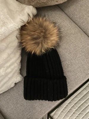 Svart mössa med pälsboll från Rockandblue - Snygg svart ribbstickad mössa från Rockandblue med stor fluffig pälsboll i brunt upptill. Mössan är tillverkad i ullblandning och har en bred uppvikt kant. Perfekt för dig som vill ha en cool vinterlook med extra attityd.