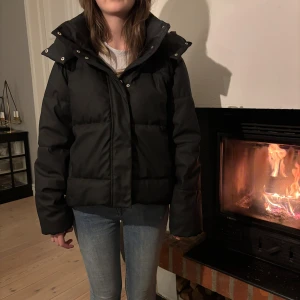 Svart pufferjacka med huva - Säljer en svart pufferjacka med stor, mysig huva och breda kanaler. Jackan har dragkedja framtill och knappar vid kragen. Perfekt för kalla dagar och har en loose passform som ger en chill vibe.