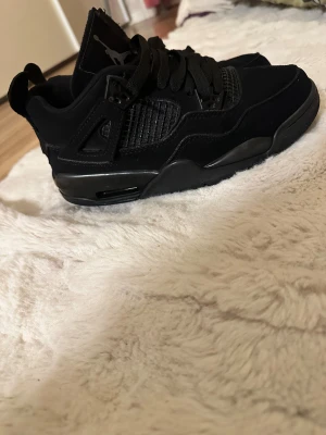 Svarta Nike Jordan 4 sneakers - Säljer ett par svarta Nike Jordan 4 sneakers med snygg meshpanel på sidorna och klassisk Jumpman-logga på plösen och sulan. Skorna har snörning, platt sula och är tillverkade i mocka och syntetmaterial. Perfekt för dig som gillar streetwear och vill ha en stilren look.