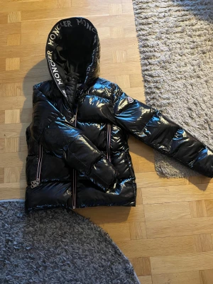 Svart Moncler pufferjacka med huva - Säljer en svart pufferjacka från Moncler med glansig finish och stor huva med Moncler-logga. Jackan har långa ärmar, två dragkedjefickor och klassisk Moncler-märke på armen. Perfekt för kalla dagar och riktigt snygg streetstil.