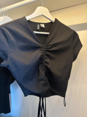 Svart croppad topp från H&M Divided - Svart ribbad croppad topp från H&M Divided med korta ärmar och snygg snörning framtill som ger en cool rynkad effekt. V-ringad hals och mjukt stretchigt material som sitter skönt. Perfekt att styla med jeans eller kjol för en trendig look.