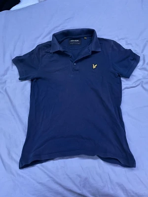 Mörkblå piké från Lyle & Scott - Snygg mörkblå pikétröja från Lyle & Scott med klassisk krage och korta ärmar. Tröjan har knappar framtill och den ikoniska gula fågeln broderad på bröstet. Tillverkad i mjuk bomull som känns skön mot huden och passar perfekt till jeans eller shorts.