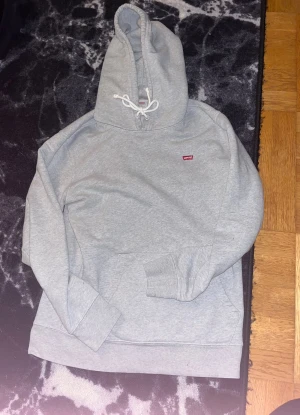 Grå hoodie från Levi's med huva - Grå hoodie från Levi's i storlek S med klassisk huva och snörning. Tröjan har en liten röd Levi's-logga på bröstet och en stor magficka framtill. Mjuk bomullskänsla och ribbade muddar vid ärmslut och nederkant. Perfekt för chill dagar.