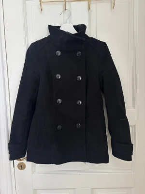 Svart skepparkavaj från H&M - Snygg svart skepparkavaj från H&M i storlek 36. Jackan har dubbelknäppning med sex knappar framtill, hög krage och två fickor. Materialet är en blandning av ull och polyester, vilket gör den både varm och stilren. Perfekt för dig som gillar klassisk look.