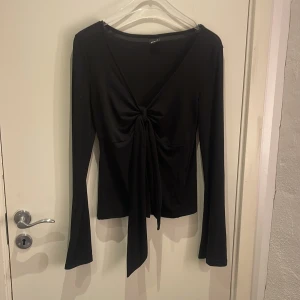 Svart långärmad topp med knyt Gina Tricot - Svart långärmad topp från Gina Tricot med djup v-ringning och snygg knytning framtill. Toppen har en figurnära passform och är tillverkad i ett mjukt, stretchigt material som känns skönt mot huden. Perfekt för att lyfta vilken outfit som helst.