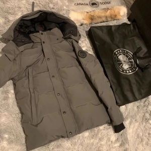 Grå dunjacka från Canada Goose - Säljer en grå dunjacka från Canada Goose med svart innerfoder och avtagbar pälskant till huvan. Jackan har lång ärm, quiltad design, knappar och dragkedja framtill samt logotyp på ärmen. Perfekt för kalla vinterdagar och har en clean, modern look.