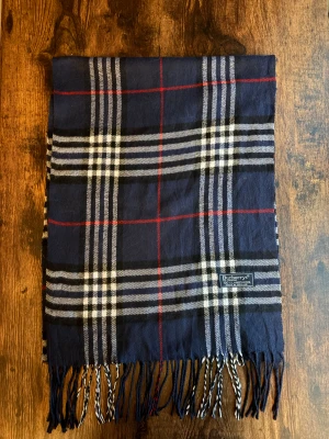 Burberry Halsduk - Klassisk Burberrys-halsduk i 100% lammull, tillverkad i England. Mjukt och varmt material i ikoniskt rutmönster. Passar både dam och herr. I mycket bra skick utan fläckar eller hål – perfekt för höst och vinter. 