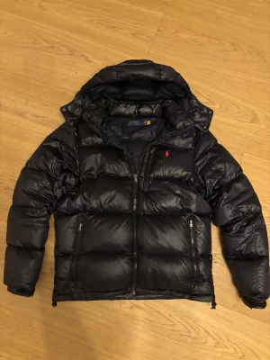 Svart pufferjacka från Polo Ralph Lauren - Svart pufferjacka från Polo Ralph Lauren med huva och dragkedja framtill. Jackan har två sidofickor med dragkedja och ett rött Polo-logo broderat på bröstet. Materialet är glansigt och jackan är vadderad för extra värme.