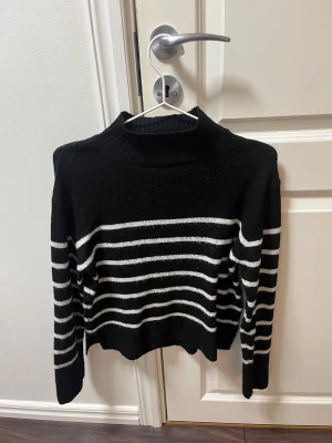 Svart och vit randig tröja  - Snygg svart polotröja med vita horisontella ränder. Tröjan har en hög krage och är långärmad, perfekt för kyliga dagar. Materialet är stickat och ger en mjuk och skön känsla. Passar dig som gillar klassisk stil med en modern twist.