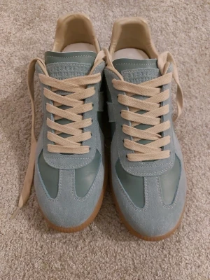 Maison Margiela blå sneakers - Maison Margiela sneakers i ljusblå mocka och skinn med beige snörning och gummisula. Klassisk rund tå och retroinspirerad design. Kommer med originalbox och dustbag. Perfekta för dig som vill ha något unikt och stilrent.