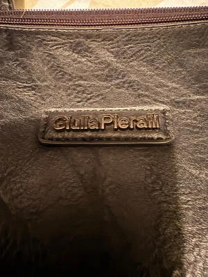 Svart axelväska från Giulia Pieralli - Snygg svart axelväska från Giulia Pieralli i skinnimitation med praktiska fack och dragkedja upptill. Väskan har långa axelremmar och rymligt innerfoder med extra fickor för småsaker. Perfekt för dig som vill ha både stil och funktion.